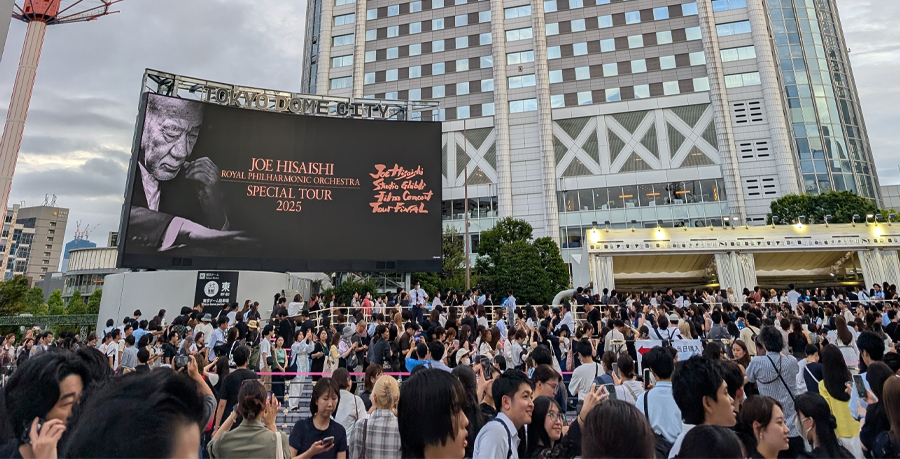 Queues of people outside Tokyo Dome with a billboard above them advertising the Joe Hisaishi and RPO Studio Ghibli music film tour